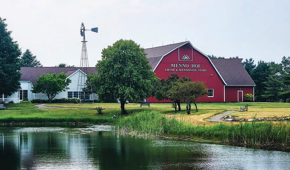 Menno-Hof tells the story of the Anabaptists. Image of red barn flanked by a white house with pond in front