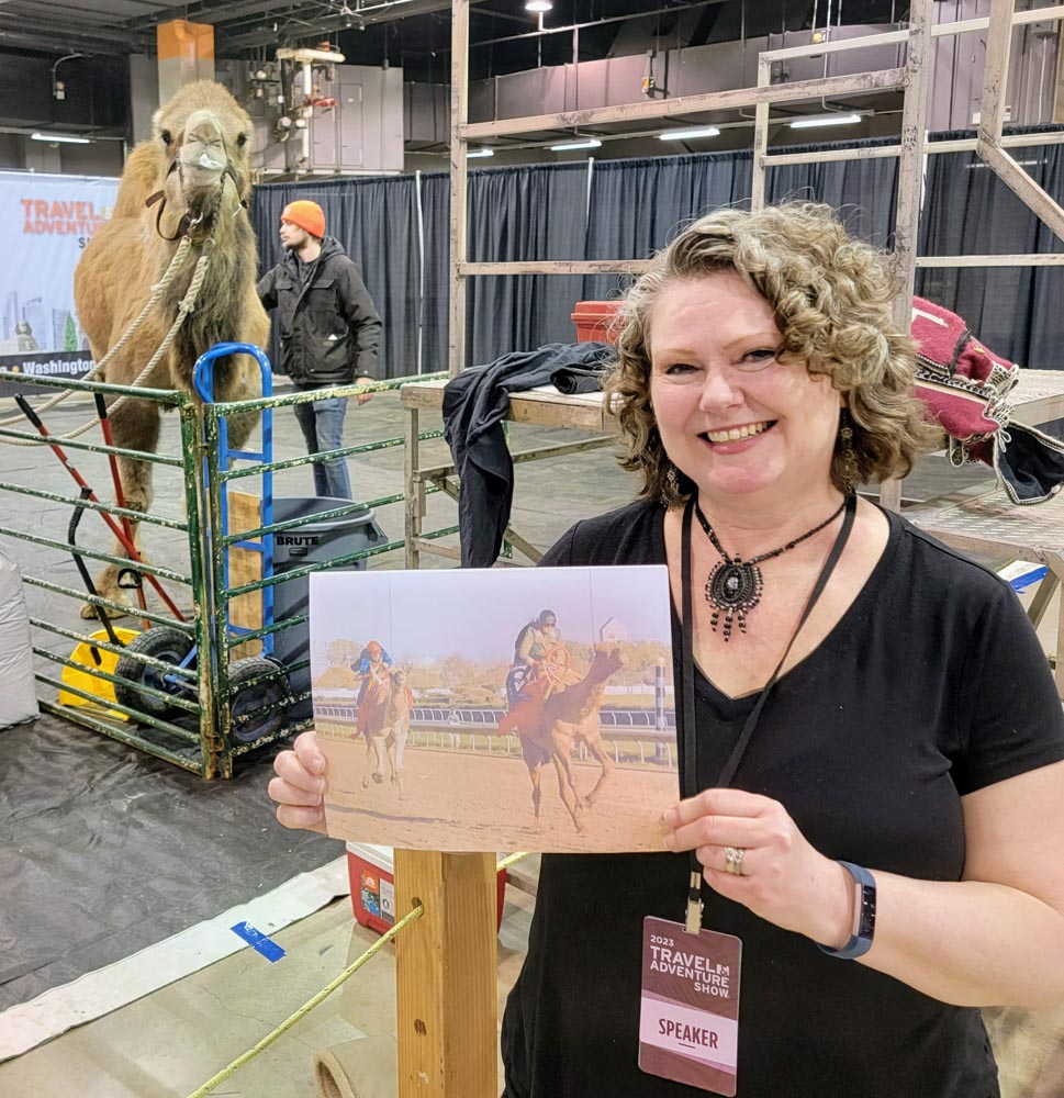 Theresa holding a picture of herself riding a camel in front of a camel